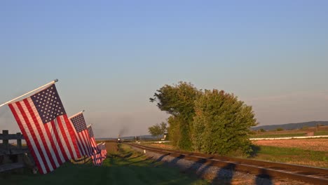 Ein-Blick-Auf-Eine-Reihe-Sanft-Wehender-Amerikanischer-Flaggen-Auf-Einem-Zaun-Neben-Ackerland,-Während-Sich-Am-Späten-Nachmittag-Ein-Dampfpersonenzug-Nähert