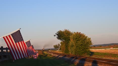 A-View-of-a-Line-of-Gently-Waving-American-Flag-on-a-Fence-by-Farmlands-as-a-Steam-Passenger-Train-Approaches-in-Late-Afternoon