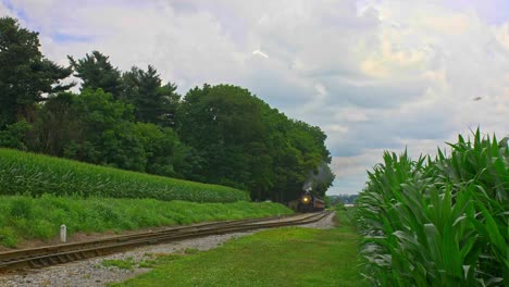 A-Front-View-of-an-Antique-Steam-Passenger-Train-Approaching-Traveling-Thru-Farmlands-and-Corn-Fields-Blowing-Smoke-on-a-Sunny-Summer-Day