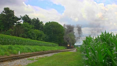 A-Front-View-of-an-Antique-Steam-Passenger-Train-Pulling-out-of-a-Stop-and-Traveling-Thru-Farmlands-and-Corn-Fields-Blowing-Black-Smoke-on-a-Sunny-Summer-Day