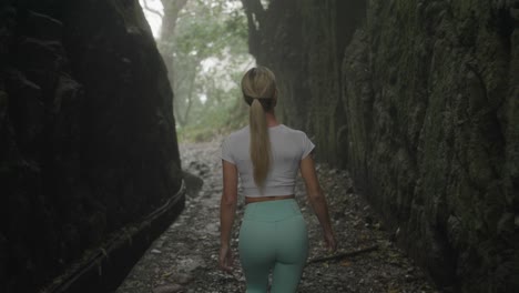 Fit-athletic-blond-woman-walking-through-misty-canyon-with-levada-waterway,-behind-shot