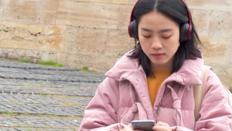 Pretty-asian-girl-using-headphones-to-listen-to-music-while-send-messages-on-her-smartphone-or-mobile-in-the-street