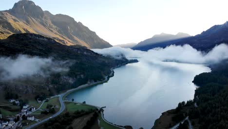 A-sunrise-aerial-flyover-up-over-Lake-Sils-in-Maloja,-Switzerland-with-a-view-of-the-peaks-of-Engadin-and-clouds-moving-over-the-water
