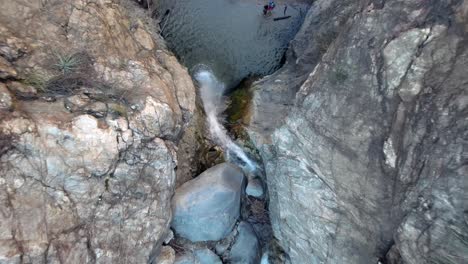Topdown-Eaton-Canyon-Falls,-drone-ascending-motion-revealing-water-flowing-down-Rocky-Cliff-Waterfall-with-tourists