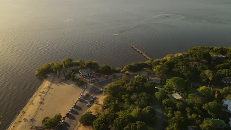 Aerial-view-showing-beautiful-coastline-with-green-trees-and-arriving-jet-ski-on-river-during-sunset---Fray-Bentos,Uruguay