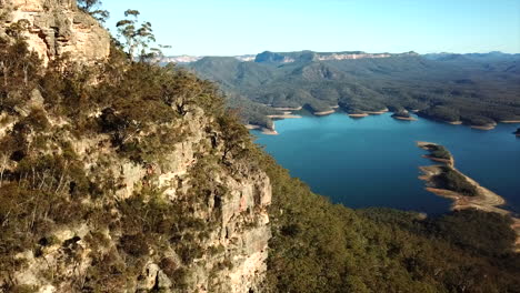 Aerial:-Drone-stationary-pan-past-a-mountain-to-a-large-beautiful-blue-lake-surrounded-by-trees-in-New-South-Wales,-Australia