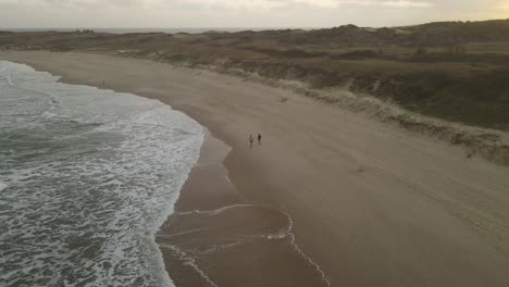 Drone-flight-following-two-people-on-walk-at-sandy-beach-during-sunset-time---Beautiful-landscape-and-atmosphere-in-the-evening---Uruguay,South-America