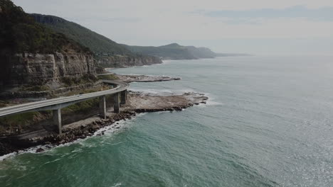 Aerial:-Drone-descending-as-a-car-drives-across-a-highway-bridge-next-to-the-ocean-in-New-South-Wales-Australia
