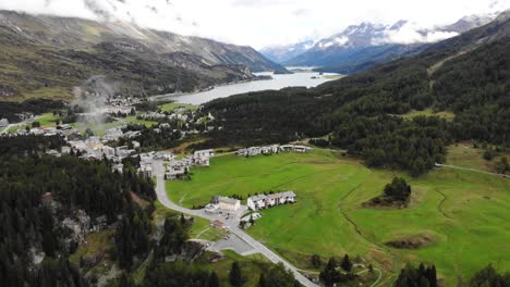 Aerial-flyover-with-pan-down-view-from-lake-Sils-down-to-the-turns-of-Maloja-Pass-in-Engadin,-Switzerland-with-cars-driving-up-and-down-on-the-road-surrounded-by-trees