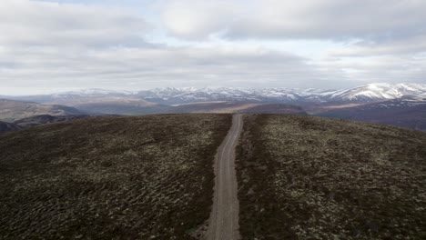 Cinematic-aerial-drone-footage-flying-up-and-over-wild-heather,-grouse-moorland-and-lichen-heath-to-reveal-a-mountain-landscape-with-patches-of-snow-and-clouds