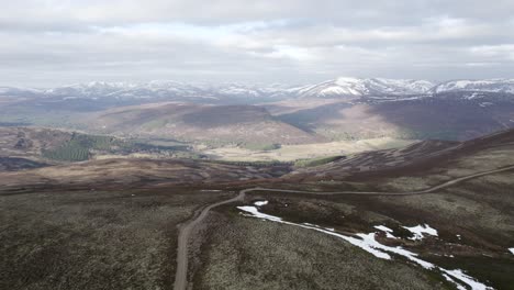 Cinematic-aerial-drone-descending-towards-a-4x4-track-and-heather-moorland-and-lichen-heath-facing-out-towards-a-mountain-landscape-with-patches-of-snow-and-clouds