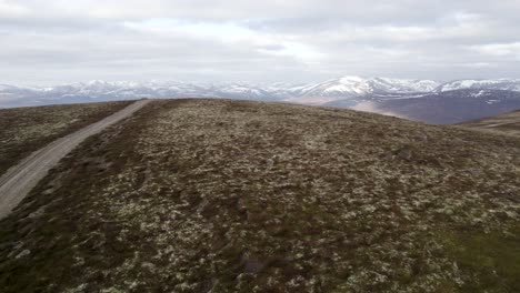 Cinematic-aerial-drone-footage-flying-fast-over-wild-heather,-grouse-moorland-and-lichen-heath-to-reveal-a-mountain-landscape-with-patches-of-snow-and-clouds