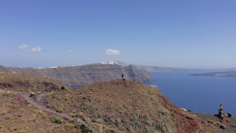 Aerial:-Flying-over-one-man-standing-at-the-top-of-hill-in-Santorini,-Greece