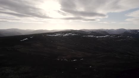 Cinematic-aerial-drone-footage-rising-above-a-mountain-landscape-and-heather-moorland-with-patches-of-snow-as-the-sun-begins-to-set-behind-the-clouds