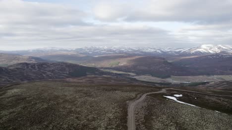 Cinematic-aerial-drone-footage-flying-over-wild-heather,-grouse-moorland-and-lichen-heath-to-reveal-a-mountain-landscape-with-patches-of-snow-and-clouds