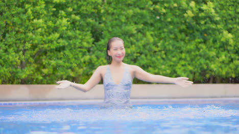 Fröhliche-Asiatische-Frau-Im-Schwimmbad,-Die-Wasser-Spielt-Und-Sprüht-Und-Mit-Einem-Lächeln-In-Die-Kamera-Schaut,-Zeitlupe
