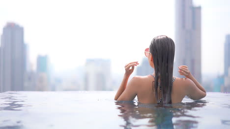 Back-view-of-sexy-woman-with-wet-hair-in-rooftop-infinity-swimming-pool-with-modern-cityscape-in-background