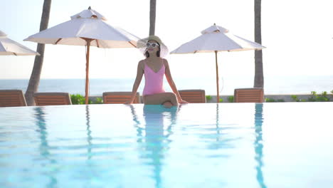 Fashionable-Asian-woman-sitting-on-infinity-swimming-pool-border-enjoying-in-warm-tropical-weather