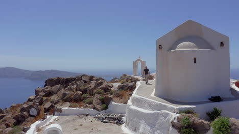 Aerial:-A-man-walks-next-to-a-church-on-a-Greek-island-on-a-sunny-day