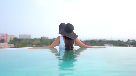Back-view-of-stylish-woman-with-floppy-summer-hat-and-swimsuit-in-swimming-pool-with-cityscape-in-background