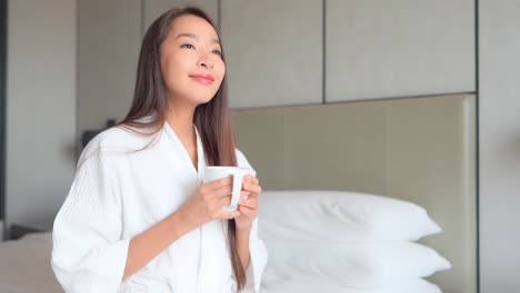 Beautiful-asian-woman-in-bathrobe-holding-cup-of-hot-coffee-or-tea-on-bed-at-sunny-bright-morning