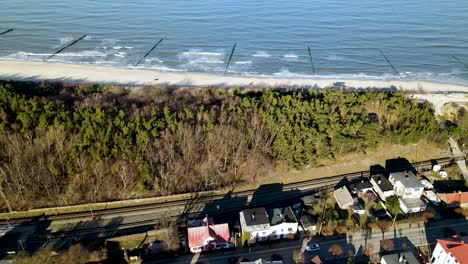 Big-waves-gently-roll-onto-the-white-beach-with-the-long-shadows-of-the-tall-trees-while-several-cars-drive-on-the-main-road-in-the-village-of-Kuznica