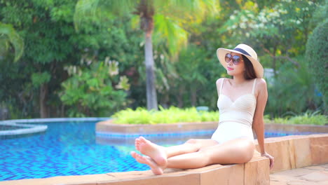 Old-fashion-exotic-woman-sitting-by-the-pool-in-swimsuit-and-floppy-hat-tropical-destination-holiday,-full-frame