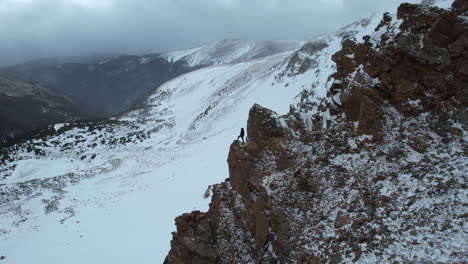 Aerial-View-of-Lonely-Man-Standing-on-Top-of-Rocky-Cliff-Above-Snow-Capped-Mountain,-Colorado-Mines-Peak,-Drone-Shot