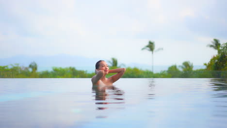 Exotic-Woman-on-Tropical-Destination-Enjoying-in-Pool-With-Green-Landscape-in-Background,-Fixing-Wet-Hair,-Slow-Motion