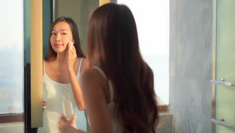 Schöne-Asiatische-Frau,-Die-Vor-Dem-Badezimmerspiegel-Ein-Anti-Aging-Hautprodukt-Auf-Das-Gesicht-Aufträgt