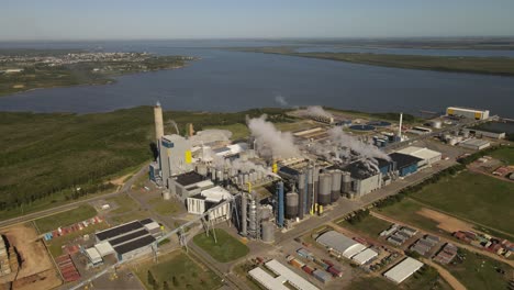 Aerial-view-of-paper-mill-factory-with-smoky-chimneys-in-front-to-River-in-Fray-Bentos,Uruguay---Environmental-pollution-of-earth