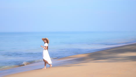 A-young-woman-wearing-a-flowing-white-sundress-and-straw-sun-hat-walks-along-the-beach-and-into-the-incoming-surf