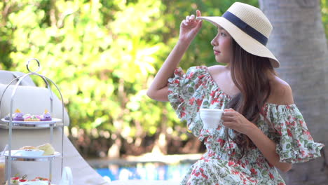 Beautiful-Asian-woman-in-dress-and-floppy-hat-sitting-in-tree-shade-with-cup-of-drink-by-the-pool-of-luxury-tropical-resort