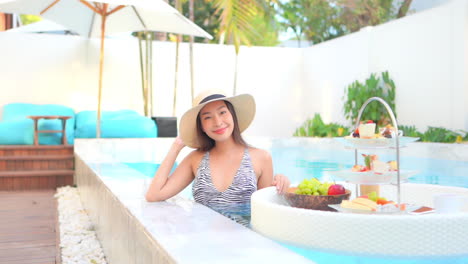 Beautiful-Classy-Asian-Woman-With-Breakfast-Floating-Plate-For-Two-in-Swimming-Pool-on-Tropical-Holiday,-Slow-Motion