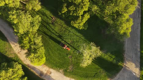 Aerial-top-down-shot-of-tractor-cutting-grass-in-park-during-sunny-day-in-the-evening---Idyllic-landscape-with-green-trees-in-summer-and-sandy-path