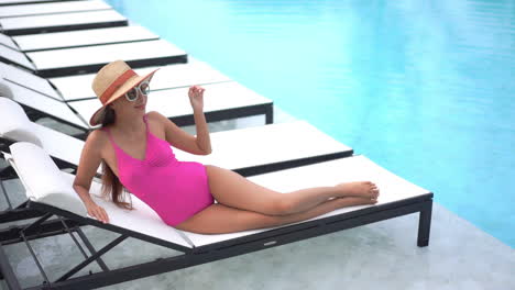 A-young-woman-in-a-pink-one-piece-swimming-suit-and-huge-straw-sun-hat-reclines-on-a-poolside-chaise-lounger