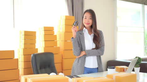 A-savvy-businesswoman-holding-a-credit-card-stands-in-front-of-multiple-stacks-of-boxes-ready-for-shipping