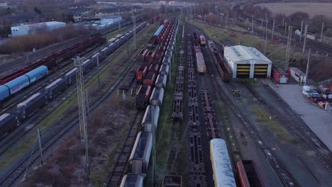 Sunrise-aerial-view-of-long-railroad-tracks-with-heavy-diesel-locomotive-carriages-and-cargo-container-yard-forward-tilt-down