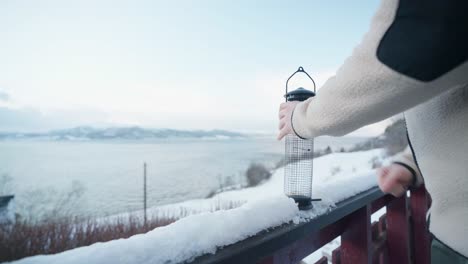 Man-Filling-Up-The-Bird-Feeder-With-Seeds-On-Snowy-Rail-Of-Fence