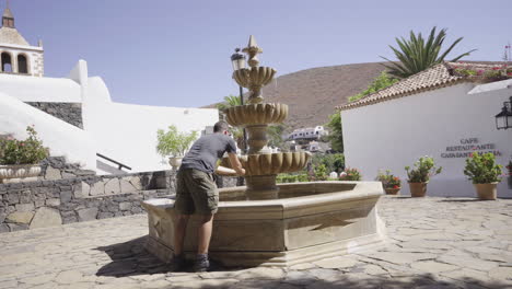 Junger-Reiserucksacktourist-Auf-Der-Kanarischen-Insel-Fuerteventura-Spanien,-Der-Aus-Einem-Wasserbrunnen-Im-Zentrum-Einer-Traditionellen-Steinstadt-In-Den-Wüstenbergen-Trinkt