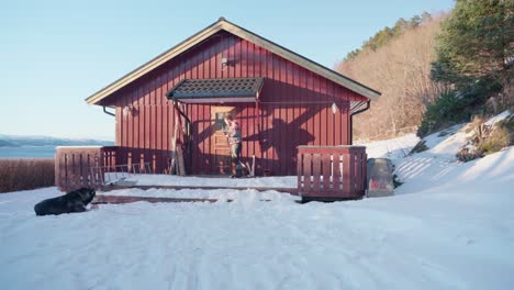 Kaukasischer-Kerl-Kommt-Aus-Der-Holzhütte,-Alaskischer-Malamute,-Der-Im-Schneebedeckten-Boden-Liegt
