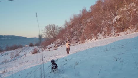 Man-Walking-His-Dog-With-A-Rope-On-Its-Neck-On-A-Snow-Covered-Hillside