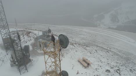Lonely-Man-on-Top-of-Cell-Tower-in-Fog-at-Snow-Capped-Mountain-Summit,-Drone-Aerial-View