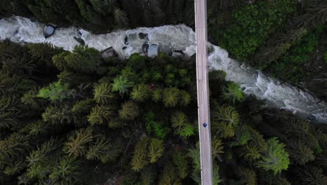 Top-down-aerial-view-following-a-hiker-walking-across-a-suspension-bridge-high-up-above-Rhone-river-valley-in-Valais,-Switzerland-at-Goms-Bridge