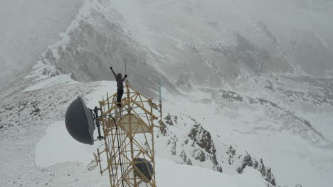 Luftaufnahme-Des-Mannes-Auf-Dem-Kommunikationsturm-Auf-Dem-Schneebedeckten-Berggipfel,-Der-Die-Arme-Hebt,-Drohnenaufnahme