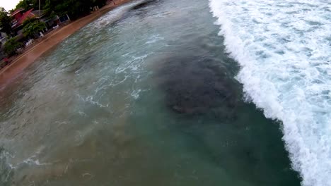 Aerial-dolly-in-lowering-on-turquoise-sea-waves-hitting-sand-shore-near-tropical-vegetation-in-Mirissa-beach,-Sri-Lanka