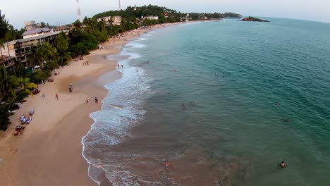Aerial-dolly-in-of-people-having-fun-in-turquoise-sea-and-sand-shore-near-hotels-and-forest-in-Mirissa-beach,-Sri-Lanka