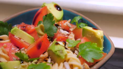 Close-up-view-of-Nutritious-pasta-salad-with-tomato,-avocado,-peppers,-nuts-and-balsamic-vinegar,-served-in-a-blue-bowl