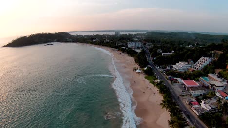 Aerial-dolly-in-over-people-relaxing-in-turquoise-sea-and-sand-shore-near-hotels-and-forest-in-Mirissa-beach-at-sunset,-Sri-Lanka