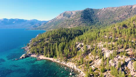 Lush-Mountains-And-Pine-Tree-Forest-In-Lake-Tahoe,-California-with-Crystal-Clear-Blue-Water-And-Beach-With-Rock-Jetty---aerial-drone-shot